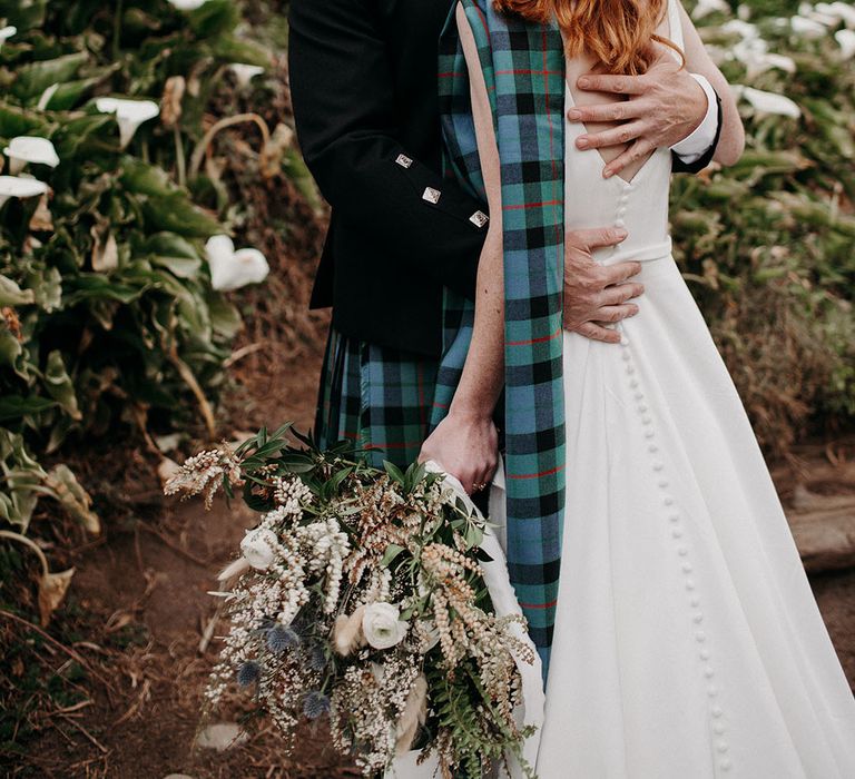 Groom embracing his bride with half up half down red hair holding an anemone and snowdrop wedding bouquet 