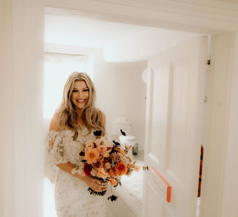 Bride in bardot wedding dress with floral applique holding white, pink and orange bridal bouquet walks out of bedroom