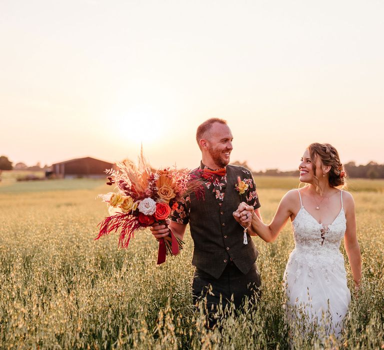 Bride in thin strap lace wedding dress holds hands with groom in short sleeve patterned shirt and bow tie with waistcoat who holds rose and dried grass wedding bouquet in field during holden hour