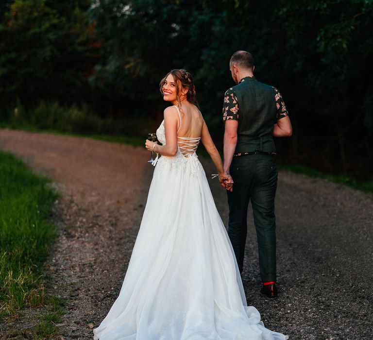 Bride in corset back wedding dress with train walks down lane holding hands with groom in short sleeve shirt and waistcoat at farm wedding