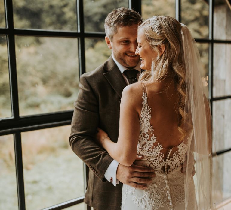 Bride in low back lace wedding dress with button detailing and veil stands with groom in brown suit by large window for rustic wedding in the Lake District