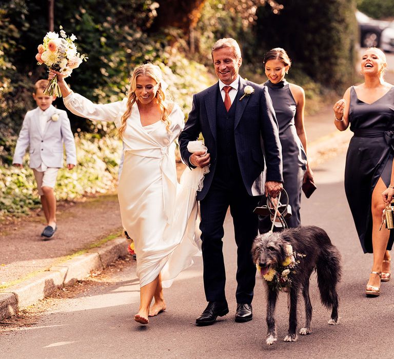 Bride in white satin dress and wrap top walks barefoot down road holding bouquet with man in blue three piece suit and dog with floral collar