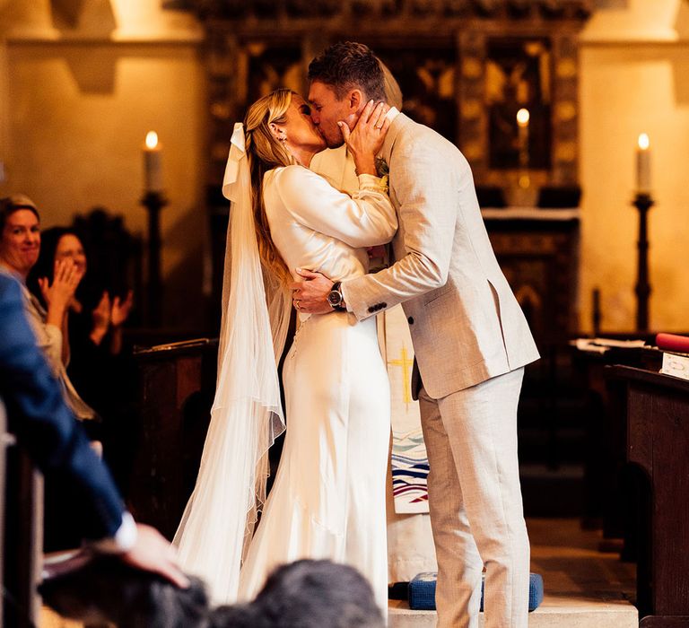 Groom in light grey suit and brown suede shoes kisses bride in satin wedding dress and wrap top wearing veil at church ceremony