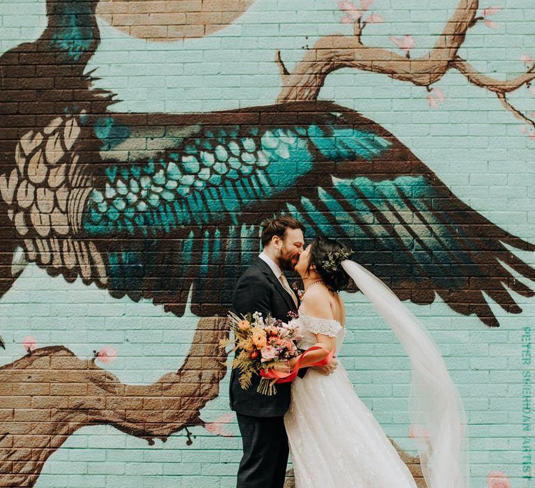 Bride in lace wedding dress with train and veil holding colourful bouquet kisses groom in check suit and brown shoes in front of large street art painting of a bird in Liverpool 