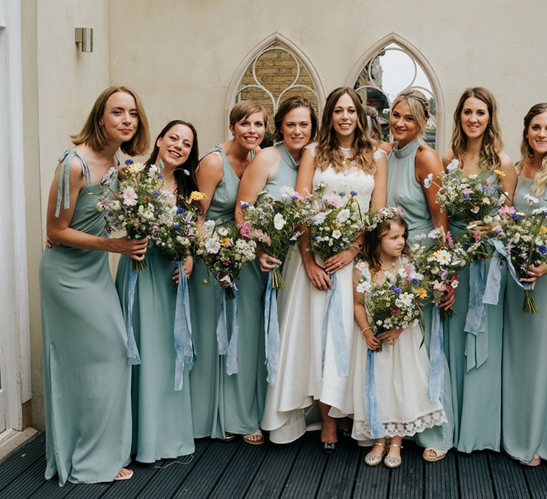 Bride with all her bridesmaids in sage green dresses with varied necklines and wild flower bouquets tied with pale blue ribbon