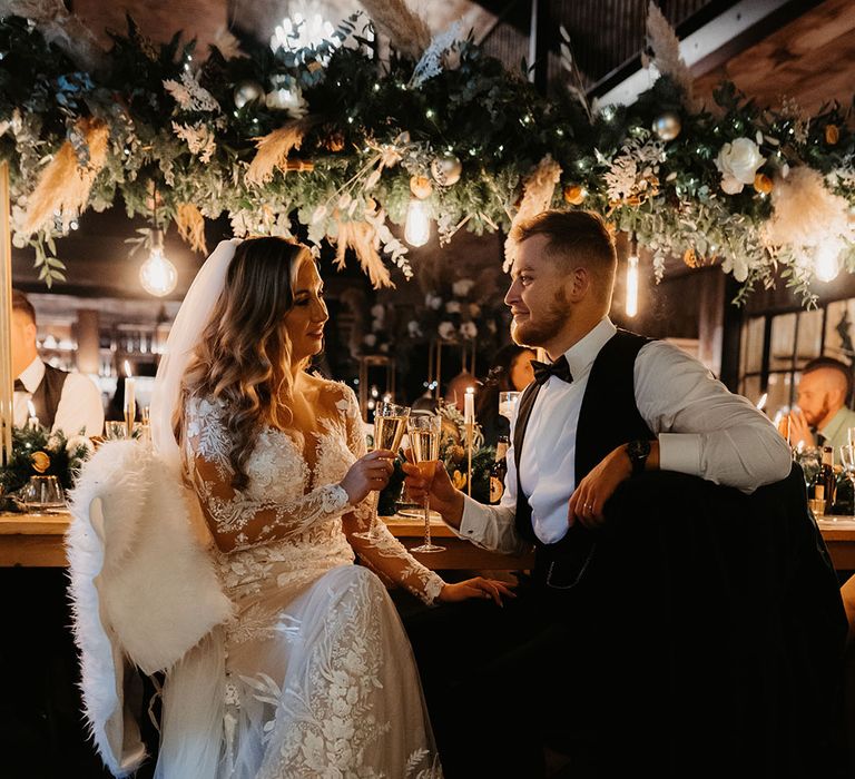 Bride and groom toast to each other with fur throws on the chairs for winter Christmas wedding 