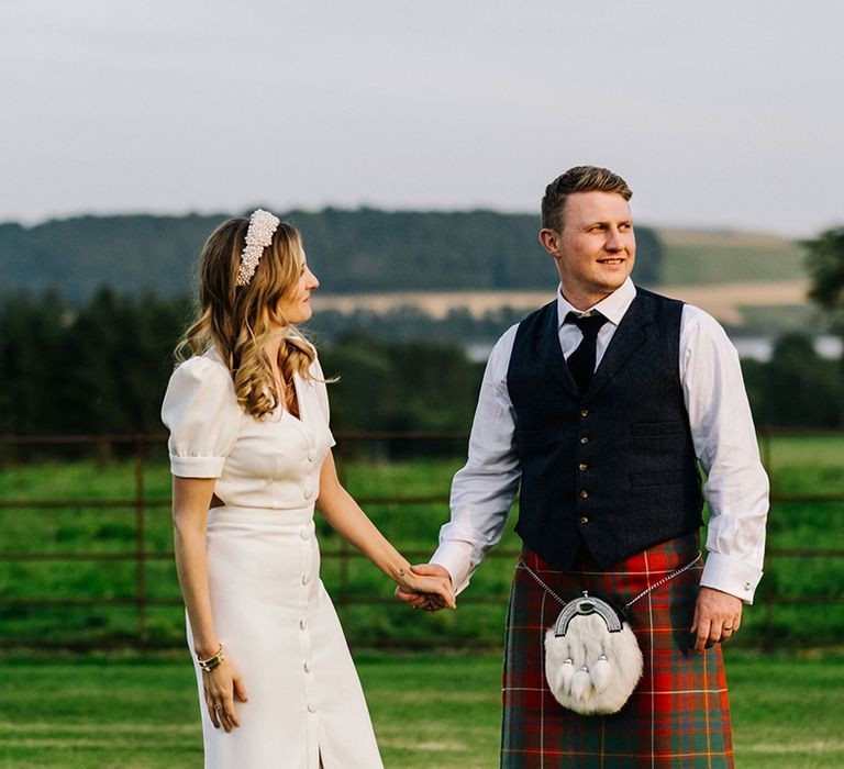 Bride in buttoned front slit puff sleeve Bon Bride wedding dress holds hands with groom in traditional kilt and blue waistcoat for country house wedding