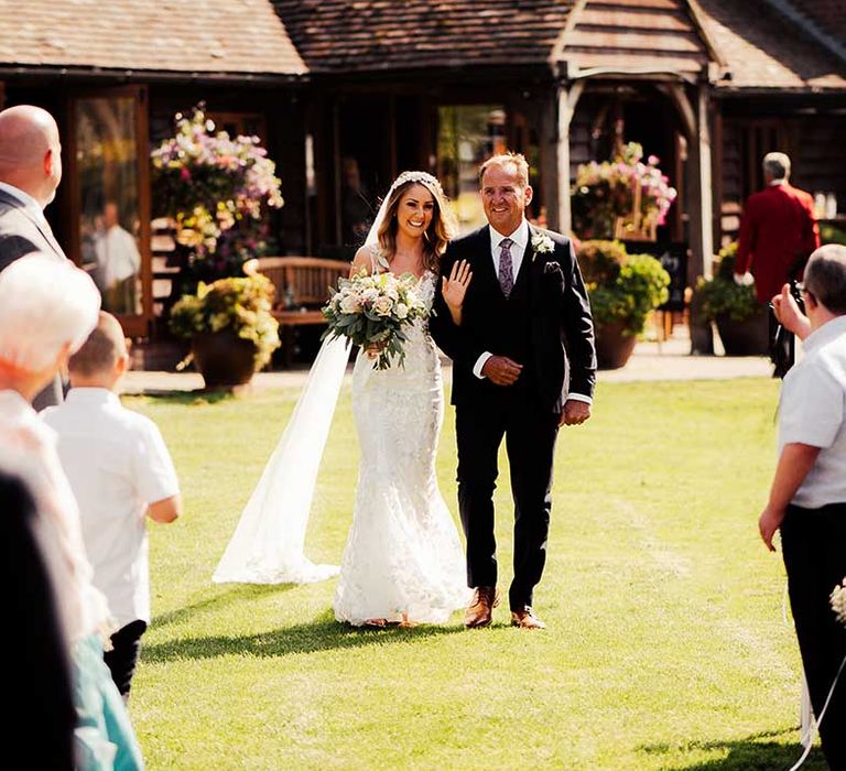 Father of the bride in dark suit, brown shoes and paisley tie walks his daughter down the aisle in lace wedding dress