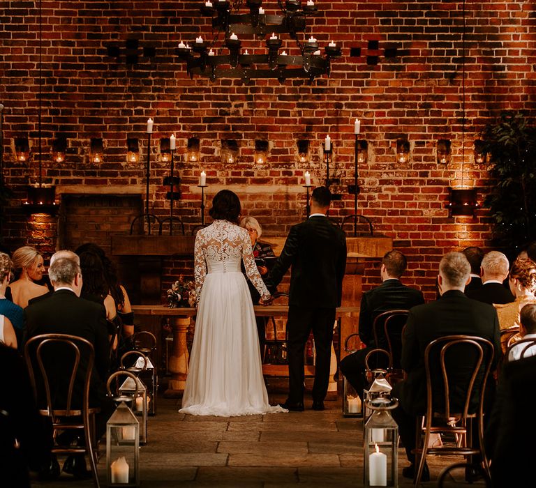 Bride in sheer lace buttoned back wedding dress and groom in black tie hold hands at the altar 