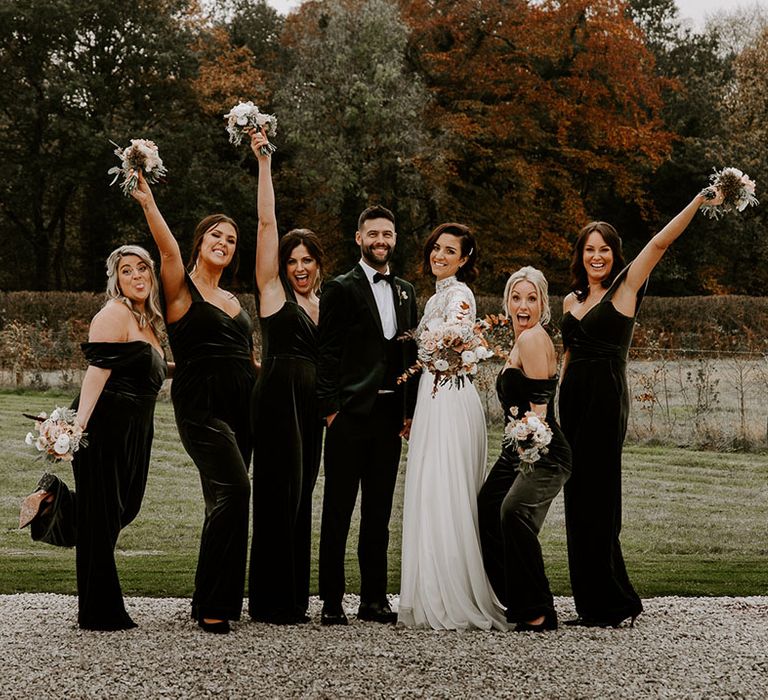 Bride and groom with bridesmaids in green velvet jumpsuits lifting their bouquets 