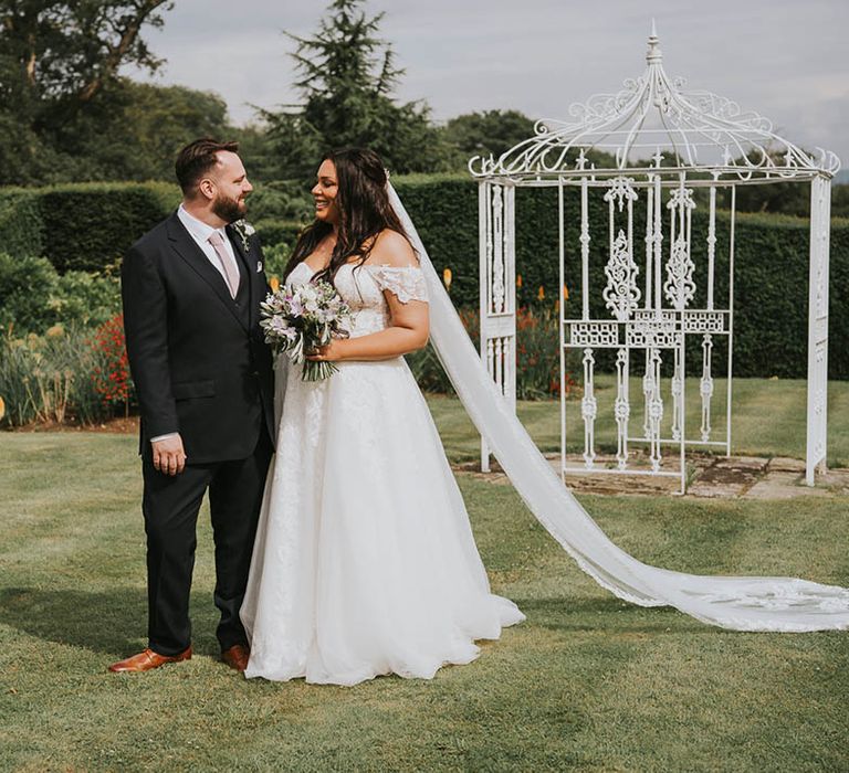 Bride wearing long lace detailed veil stands with the groom in a dark grey suit and pink tie 