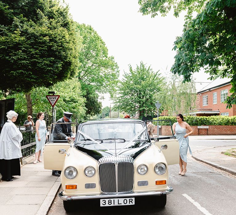 Bridesmaids in blue one shoulder wrap dresses as they help the bride get out the cream and black wedding car transport