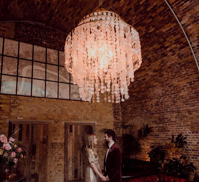 Red leather sofa and modern chandelier decorate the bride and groom's wedding venue as they hold hands under the light 