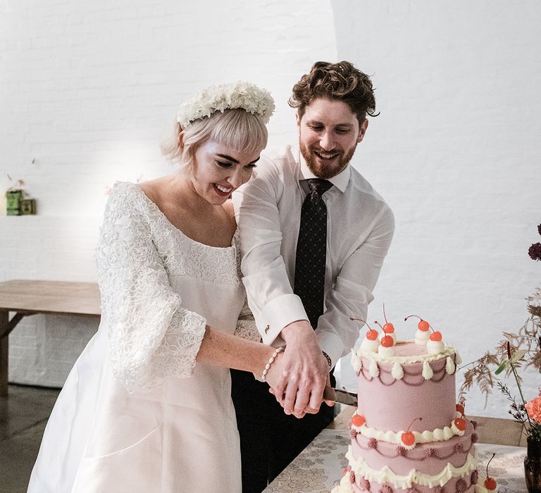 Bride in short wedding dress and groom in patterned tie cut their three tier purple and white wedding cake decorated with cherries and cupcakes