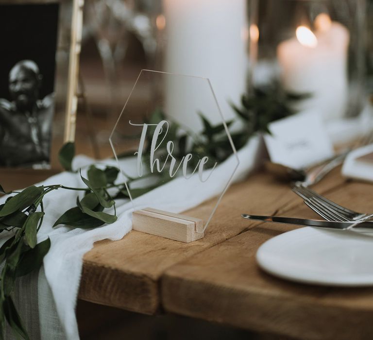 Hexagonal clear table number sign in wooden holder for classic wedding table setting with green foliage and white runners 