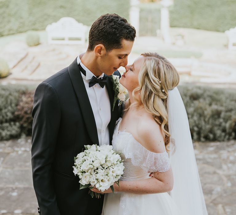 Cute bride and groom couple portrait with bride in lace off the shoulder wedding dress with pearl hair pins and white bouquet with groom in black tie 