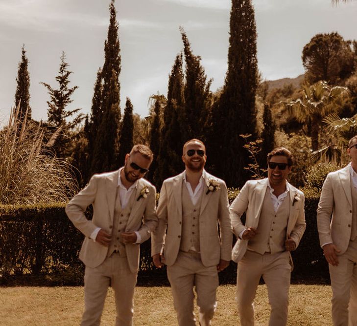 Groom walks with his groomsmen who wear sunglasses, cream three piece suits and open relaxed shirts in Spain 