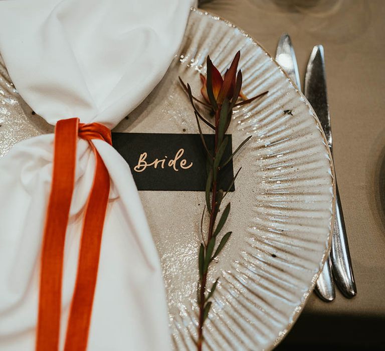 White and silver wedding plate with white napkin tied with burnt orange ribbon with black name place card and flower