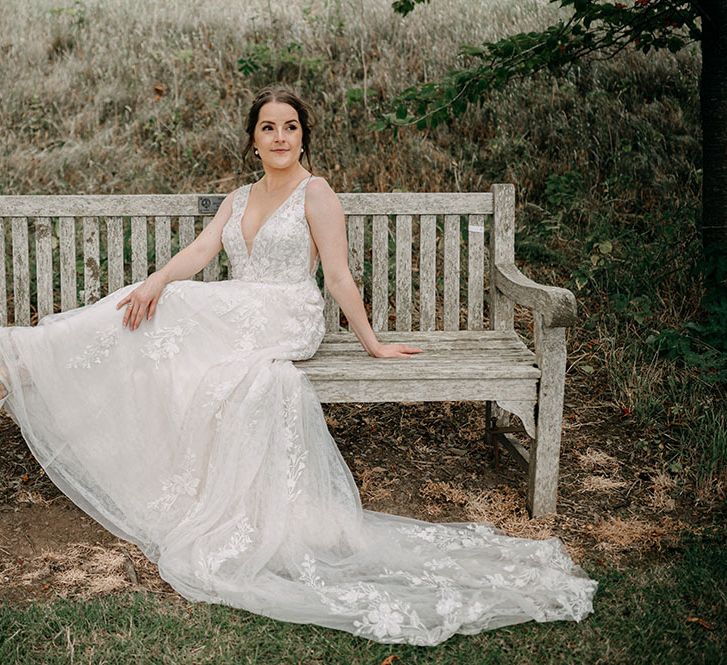 Bride wears v-neck lace wedding dress with sequin embellishment and satin pointed shoes as she sits across wooden bench outdoors