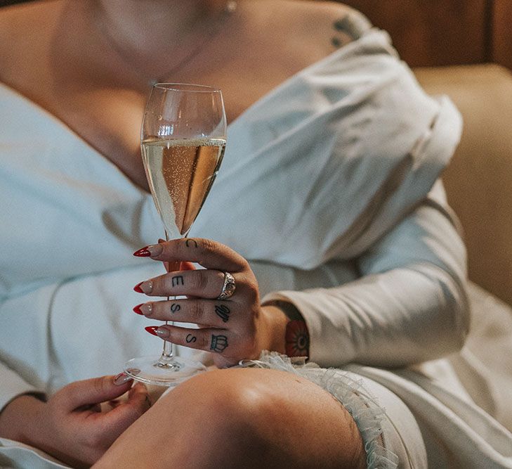 Bride wears off-the-shoulder ASOS curve wedding dress and holds champagne glass in her hands with red tip manicured fingernails 