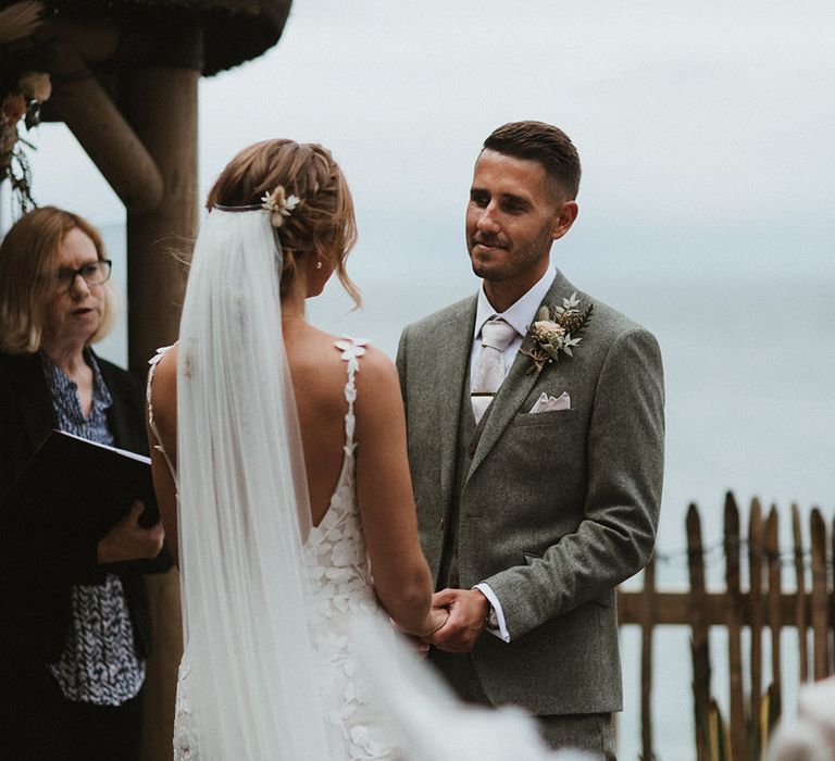 Bride wears floor-length veil pinned into her fishtail plaited hair as she and her groom say their vows during outdoor wedding ceremony