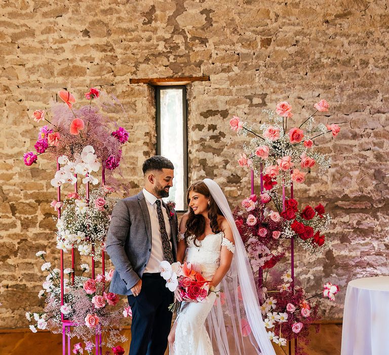 Bride wears lace wedding dress whilst stood with her groom in grey suit in front of a bright floral arch 