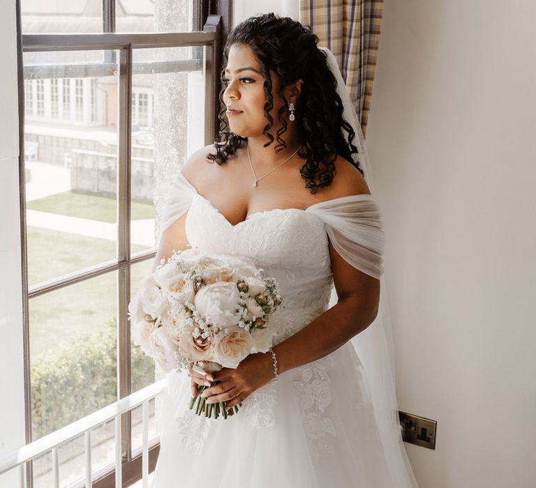 Bride wears sheer sleeved off the shoulder wedding dress with lace and floral embellishment whilst holding classic bouquet 