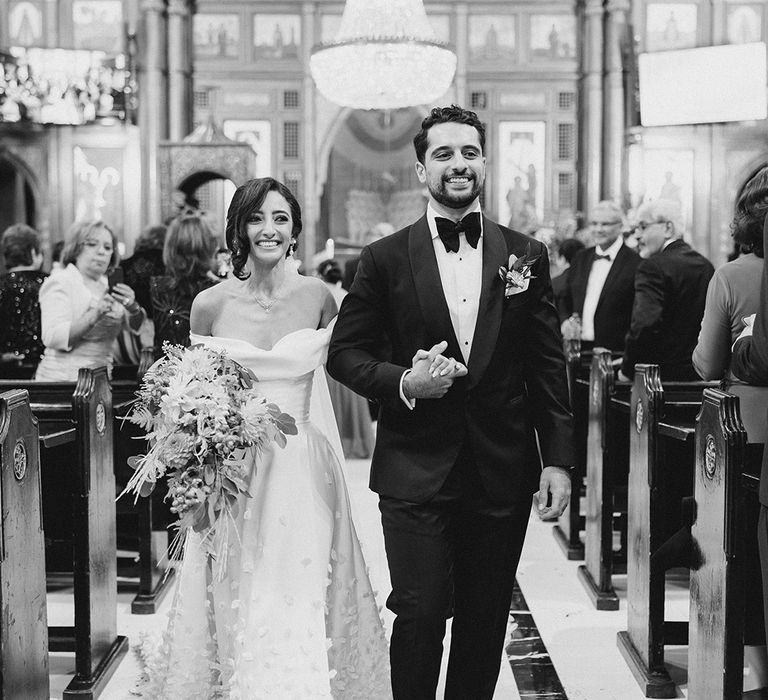 Bride in off-the-shoulder styled wedding dress walks down the aisle with her groom in black-tie 