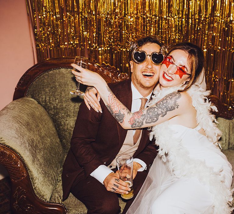 Bride & groom sit in DIY photo booth wearing fun sunglasses and gold streamers to the back