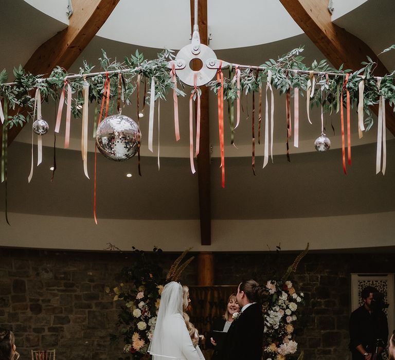 Bride in square neck long sleeve satin wedding dress with two tier veil holding hands with groom in black suit with floral boutonniere at the alter