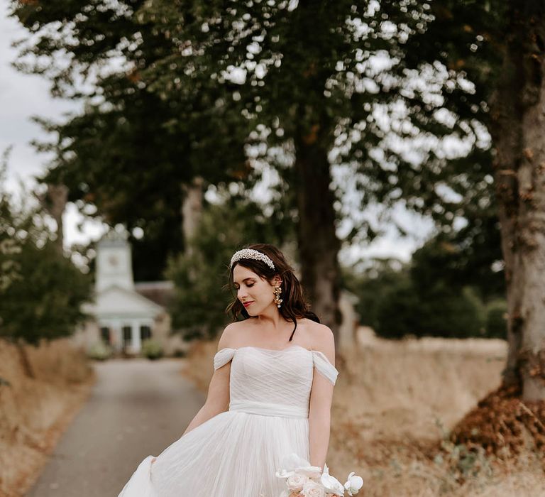 Bride in off the shoulder wedding dress wearing a pearl headband and statement earrings with neutral wedding bouquet 