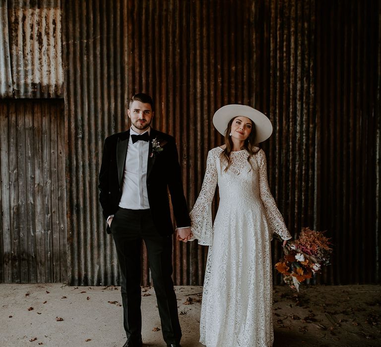 Groom in green tuxedo holding hands with the bride in a bell sleeve lace wedding dress with a white hat with dried flowers 