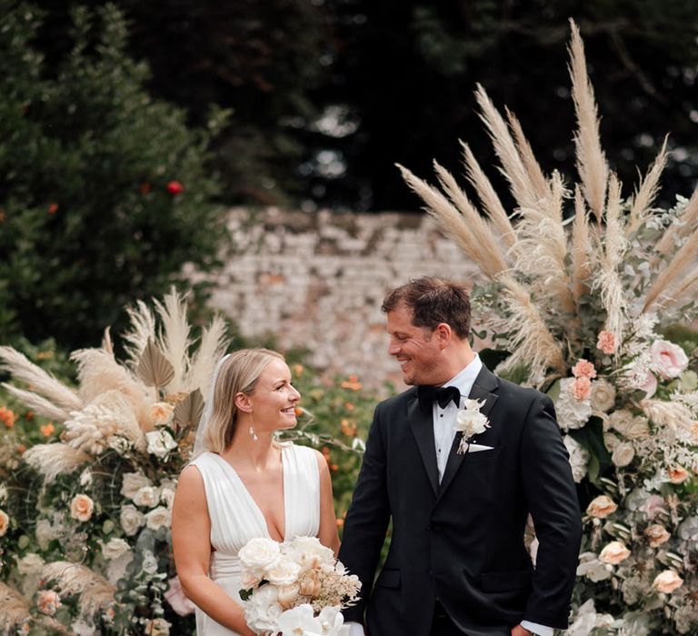 The bride and groom smile at each other standing in front of flower columns with pampas grass with pale pink and white roses 