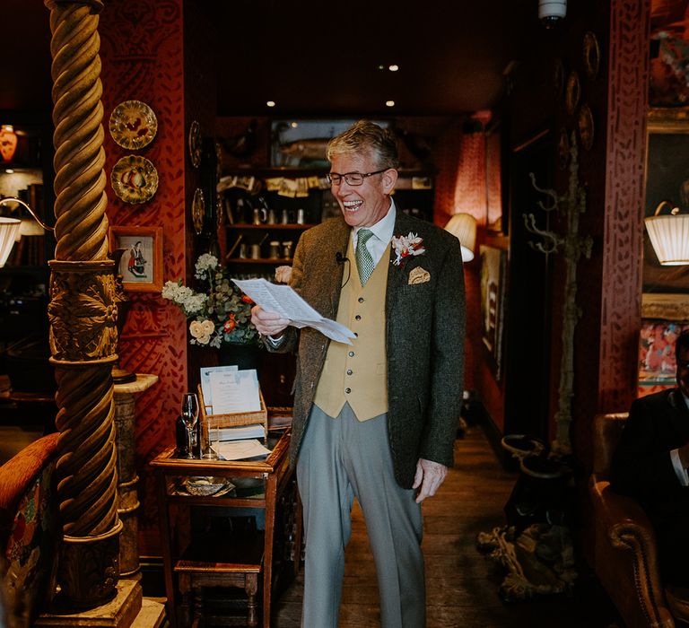 Father of the bride wearing dark grey blazer, grey trousers, yellow waistcoat, green tie and autumnal boutonniere giving a speech at intimate London wedding