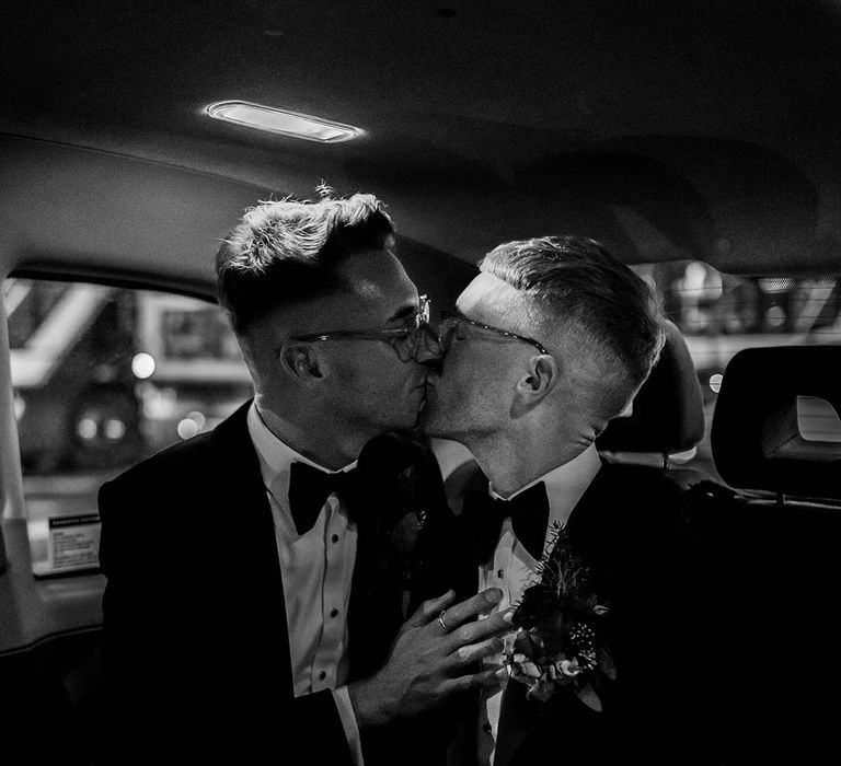 Grooms in matching velvet tuxedos with black bowties and rose and foliage boutonnieres kissing in the back of a wedding car