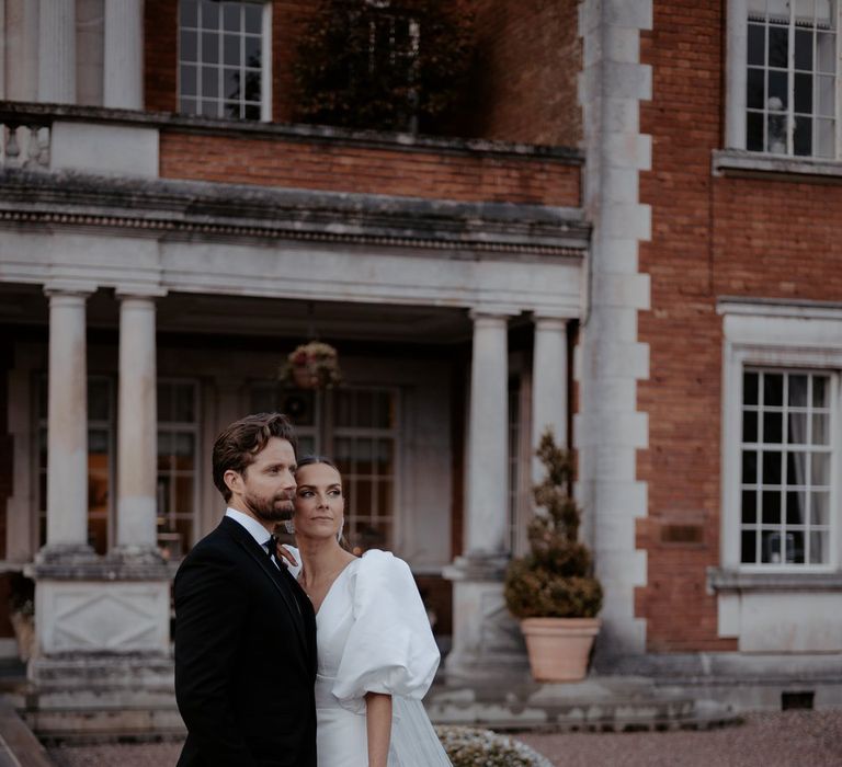 Groom wearing classic black tux with bowtie embracing bride in v neck puff sleeve wedding dress with front slit and church length veil standing in the gardens of Eaves Hall Clitheroe 