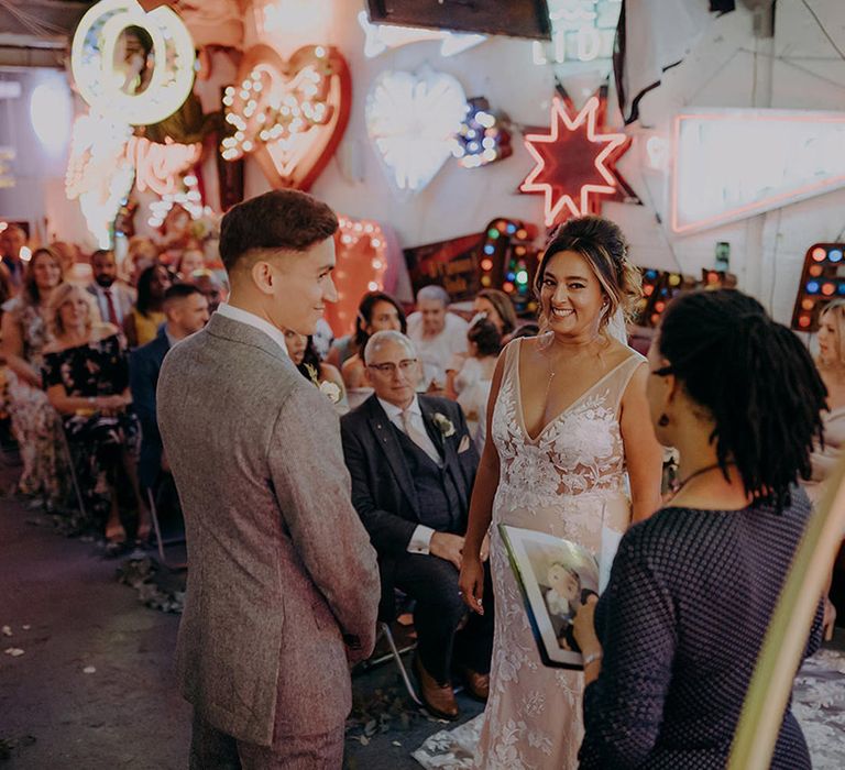 Groom in grey three piece wedding suit with yellow suit and yellow pocket square standing with bride in sleeveless plunge lace wedding dress  and cathedral length embroidered veil with celebrant in navy polka dot dress at London wedding 