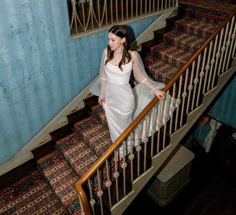 Bride in long sheer puff sleeve satin wedding dress with cowl neck walking down the stairs of St Giles House 