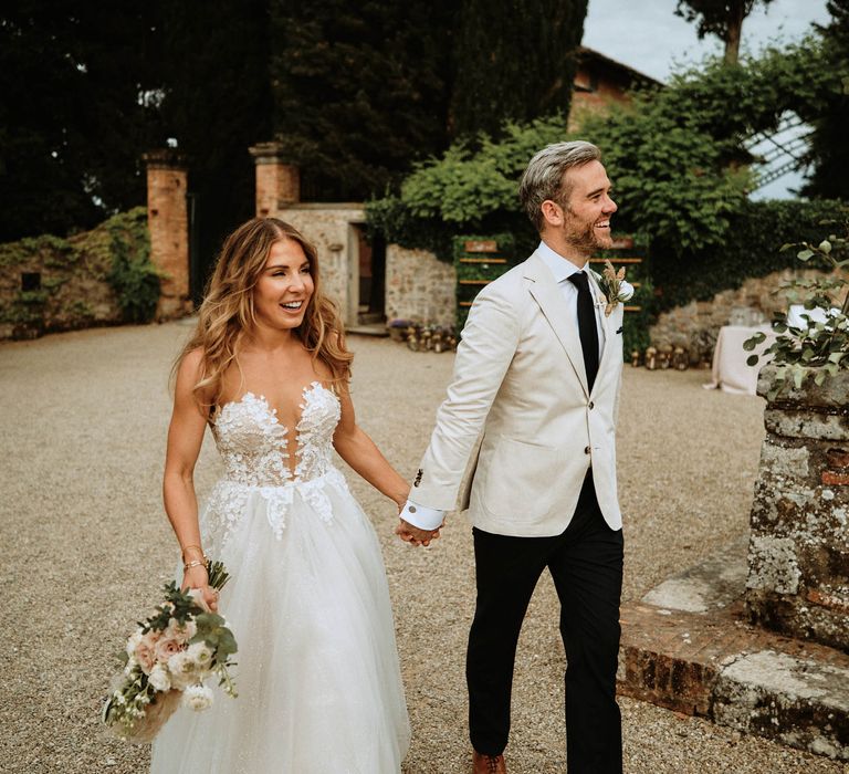 Bride and groom holding hands and smiling at italian wedding wearing a tulle wedding dress with embroidered bodice with groom in linen suit