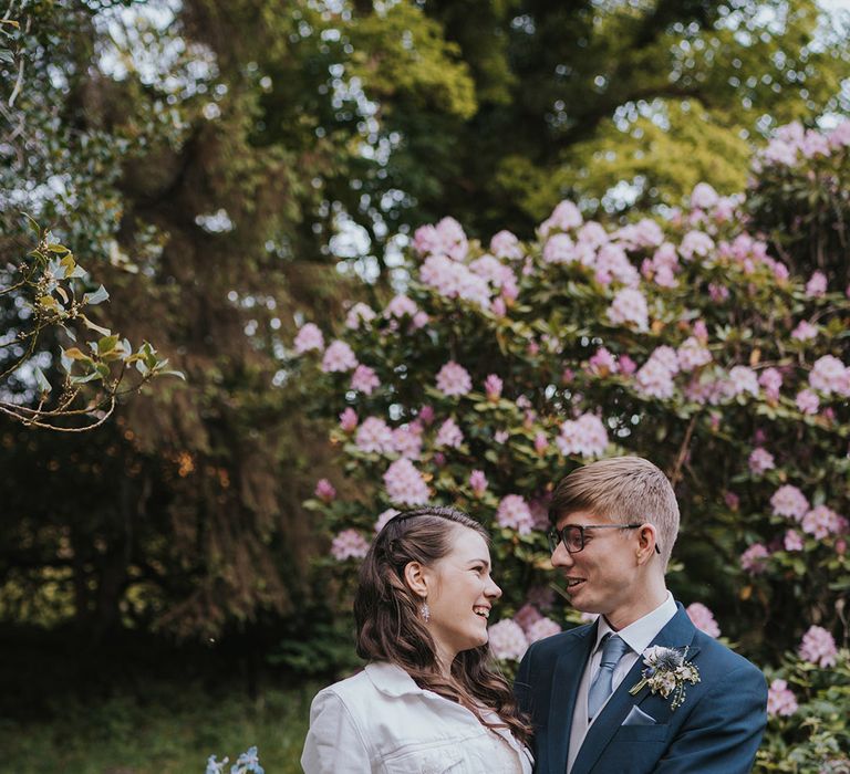 Bride in personalised white denim jacket smiling with the groom in navy suit at their woodland setting at Badgers Holt in Devon