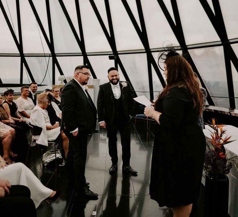 Wedding celebrant wearing black long sleeve dress with black wedding fascinator leading the wedding at The Gherkin with grooms wearing complimenting three piece black tuxedos with off white bow ties, white garden rose, lavender twig and dried foliage boutonnieres and grooms Louboutins shoes 