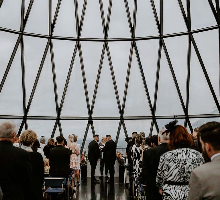 Grooms wearing three piece black tuxedos with off white bow ties and white garden rose, lavender twig and dried foliage boutonnieres doing the ring exchange at the end of the aisle at The Gherkin London 