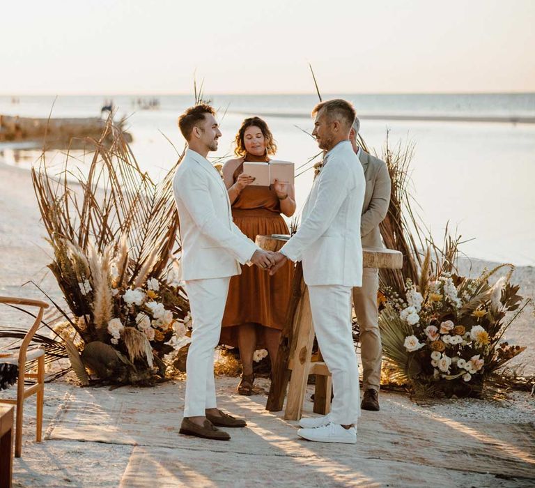 Groom in mens white wedding suit, light blue and white striped shirt, dusky orange garden rose and dried flower boutonniere and gold jewellery holding hands with groom in white blazer, light blue and white striped shirt, dusky orange garden rose and dried flower boutonniere and gold jewellery