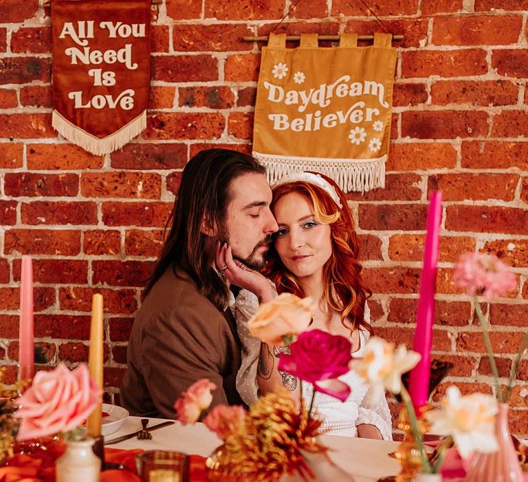 Bride in three quarter length puff sheer sleeve wedding dress with delicate polka dot overlay and square neck and white bridal headband posing with groom in brown grooms blazer at retro inspired wedding tablescape at Northern Monk Refectory Leeds