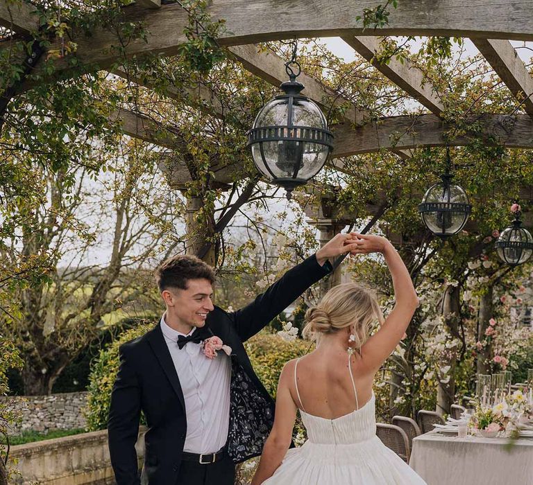 Groom in classic black tuxedo, bowtie and blush rose boutonniere spinning bride in Watters strappy sleeveless princess wedding dress by al fresco outdoor wedding tablescape 