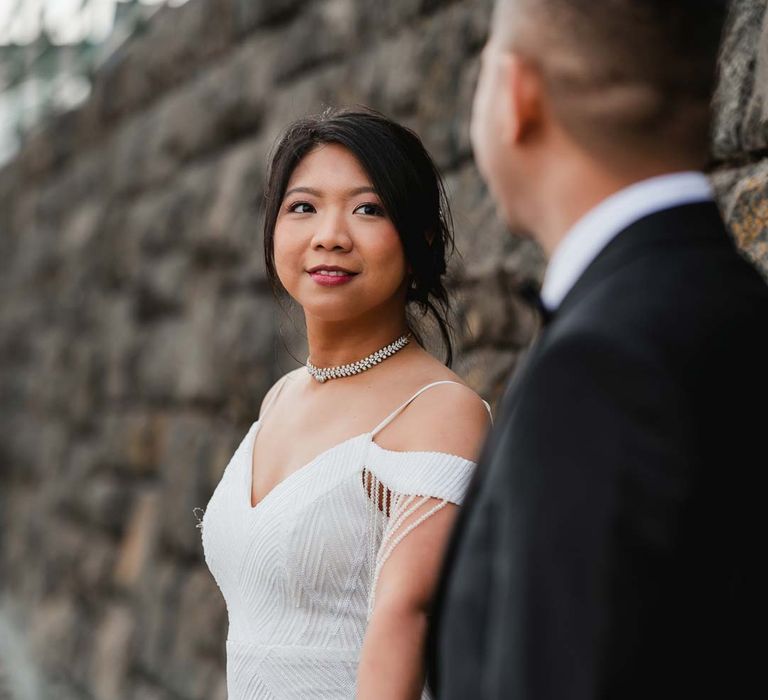 Bride in white beaded and sequined slip wedding dress with tassels on the sleeves holding hands with groom in classic black tux 