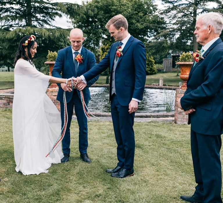 Bride in long sleeve mesh overlay wedding dress and colourful bridal flower crown doing handfasting ceremony with groom in classic navy grooms suit with black tie, grey waistcoat and colourful rose boutonniere at Templars Barn