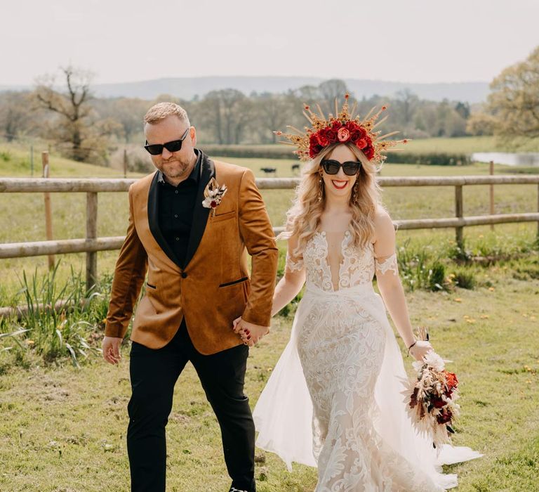 Bride in off shoulder lace wedding dress with corset style top and red rose, gold halo bridal crown and sunglasses holding red rose, pampas grass and dried flower bridal bouquet walking with groom incrushed velvet burnt orange grooms blazer and dried flower boutonniere and sunglasses at Southlands Barn wedding