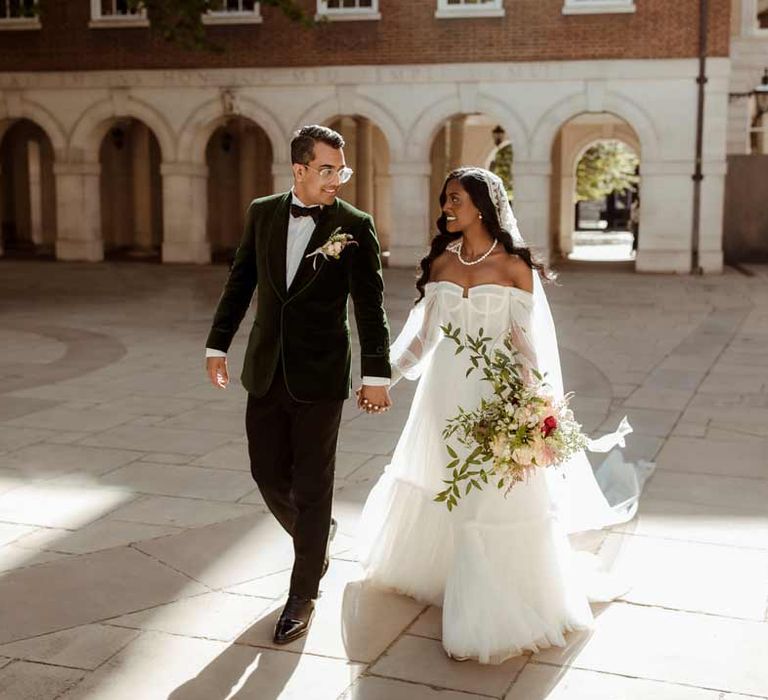 Groom in bottle green velvet grooms suit, black bowtie and chic boutonniere walking with bride in off the shoulder layered tulle wedding dress and floral embroidered cathedral-length veil at Inner Temple Hall vow renewal