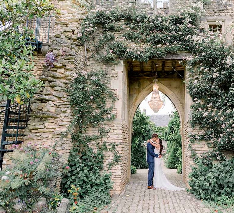 Groom in a navy suit and bride in a fitted wedding dress kissing under the crumbling arch with luxury chandelier 